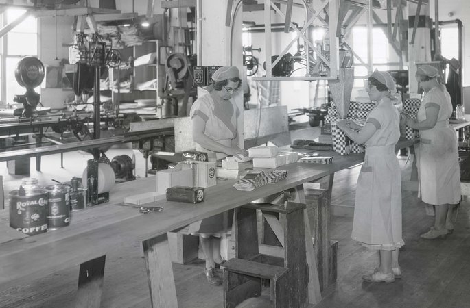 Photograph showing three unidentified women standing at a long table and filling bags of coffee, possibly Royal Club coffee. They are in a large room with machinery in the background.