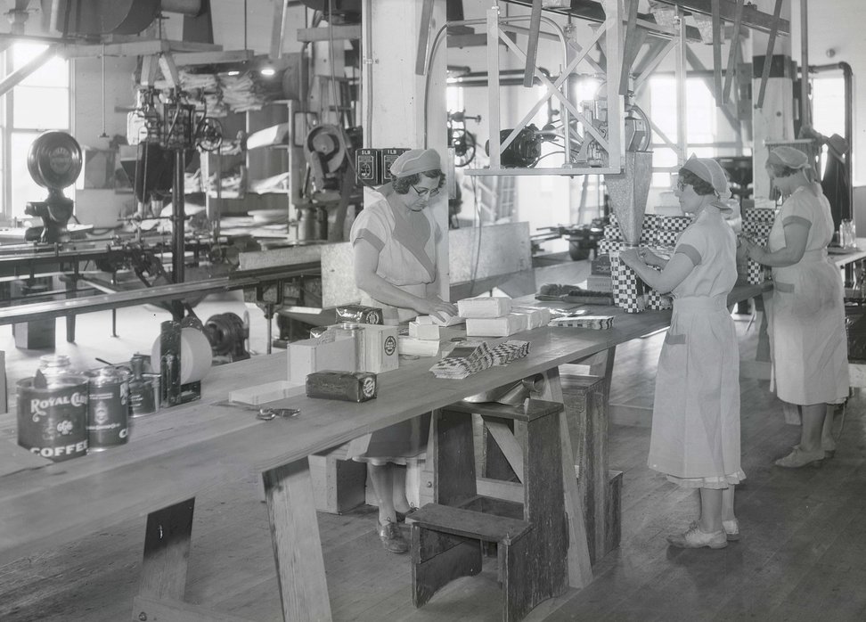 Photograph showing three unidentified women standing at a long table and filling bags of coffee, possibly Royal Club coffee. They are in a large room with machinery in the background.