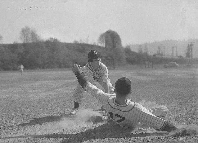 Softball player Margaret Dobson playing for the Vanport College baseball team. She leans downward towards a sliding player, most likely to tag him out. Another player can be seen in the distance, standing on the dirt field. Dobson was inducted into the Oregon Sports Hall of Fame in 1984